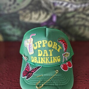 May include: Green trucker hat with the words "Support Day Drinking" in yellow. The hat features patches of a cocktail, watermelon, cherries, and a butterfly. A gold safety pin and hook are attached.