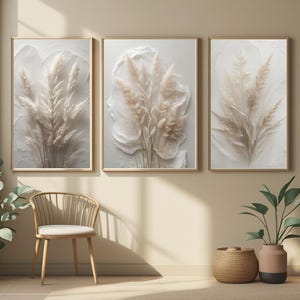 Peut inclure: Trois œuvres d'art encadrées représentant des herbes de la pampa beiges sur un fond blanc texturé. L'œuvre est dans des cadres dorés. La pièce a une palette de couleurs neutres avec une chaise en bois, un panier tressé et des plantes en pot.