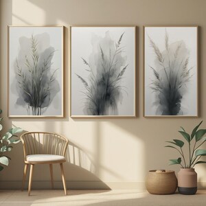 Peut inclure: Trois impressions encadrées à l'aquarelle représentant des herbes hautes abstraites dans des tons de gris et de vert. L'œuvre est exposée sur un mur beige, avec une chaise en bois, un panier tressé et des plantes en pot au premier plan.
