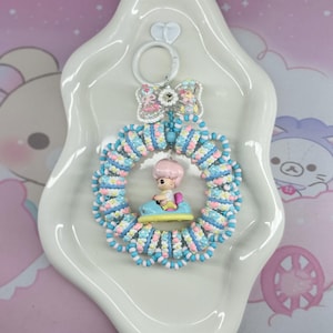 May include: A whimsical keychain featuring a miniature figure in a blue car, surrounded by a beaded wreath in pastel colors. The keychain includes a decorative bow and is set on a white, heart-shaped tray.