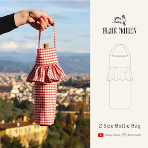 May include: A red and white gingham bottle bag with a frilled skirt design, held by a handle. The bag is designed to hold a wine bottle, with a cork visible at the top. The bag is approximately 30 cm tall.
