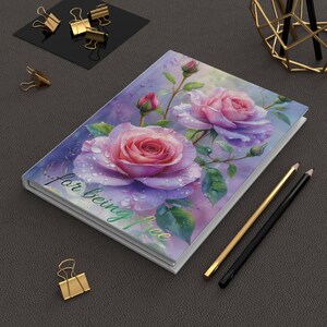 Peut inclure: Un journal à couverture rigide avec une peinture à l'aquarelle de roses roses avec des gouttelettes d'eau. La couverture porte l'inscription "...for being free" en vert. Des crayons dorés et noirs et des trombones dorés sont également visibles.