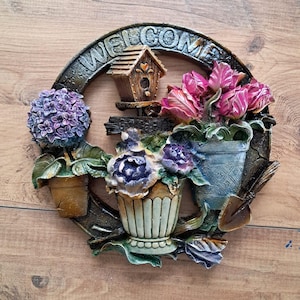 May include: A decorative wreath with the word "WELCOME" in a circular design. The wreath features a small birdhouse, potted flowers in shades of purple and pink, and a small shovel. The background is a wooden surface.