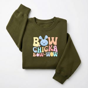 May include: An olive green sweatshirt with the text "Bow Chicka Bow-Wow" in pastel colors. The design includes a blue bunny face, flowers, and an Easter egg. The sweatshirt has a crew neck and long sleeves.