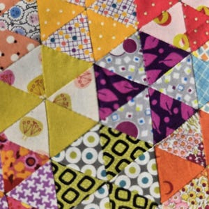 May include: Close-up of a patchwork quilt with a geometric design. The quilt features various fabrics in shades of yellow, orange, purple, pink, and green. The pattern is made up of triangles and hexagons, creating a colorful and textured surface.