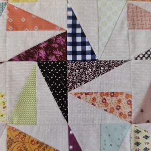 May include: Close-up of a quilt with a starburst design. The quilt features various patterned fabrics in orange, purple, black, and blue, arranged in a geometric pattern. The background is a light beige fabric.
