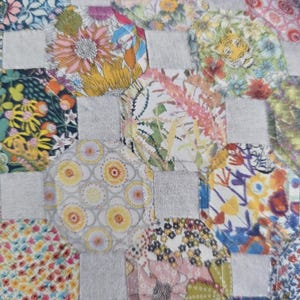 May include: Close-up of a patchwork quilt with a grid pattern. The quilt features floral fabric patches in various colors, including yellow, orange, pink, and blue, set against gray squares. The floral patterns include daisies, sunflowers, and other stylized flowers.