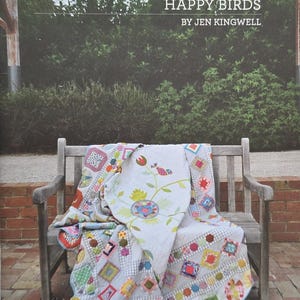 May include: A colorful quilt with a patchwork design is draped over a wooden bench. The quilt features various geometric shapes and patterns in shades of blue, green, red, and yellow. The text "HAPPY BIRDS" is visible at the top of the image.