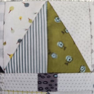 May include: Close-up of a patchwork design featuring a tree-like shape. The tree is composed of various fabrics, including white with small yellow and gray shapes, vertical blue and white stripes, and olive green with small blue floral patterns. The trunk is a dark gray with small black spots.