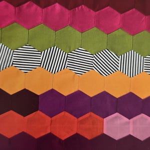 May include: A colorful patchwork textile featuring a repeating hexagonal pattern. The design incorporates various colors, including pink, green, orange, purple, and red. Some hexagons have black and white striped fabric.