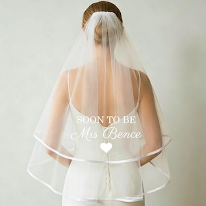 Pode incluir: Um véu de noiva branco com um design de camada dupla e uma borda de fita de cetim. O véu apresenta o texto "SOON TO BE Mrs Bence" e um coração. A noiva está usando um vestido branco.