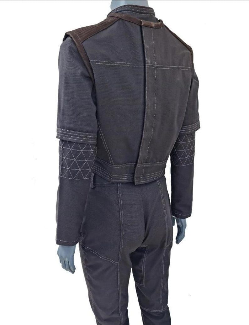 May include: Dark gray jumpsuit with brown shoulder accents and white stitching. The sleeves have geometric patterns near the elbow. The back of the jumpsuit has a vertical seam and horizontal stitching across the shoulders and waist.