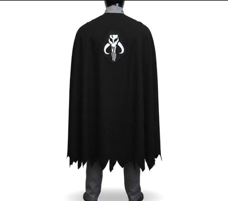 May include: A black cape featuring a white Mythosaur skull emblem centered on the upper back. The cape has a jagged, uneven hemline at the bottom and is worn over a grey collared shirt and grey trousers, covering the back of the torso.