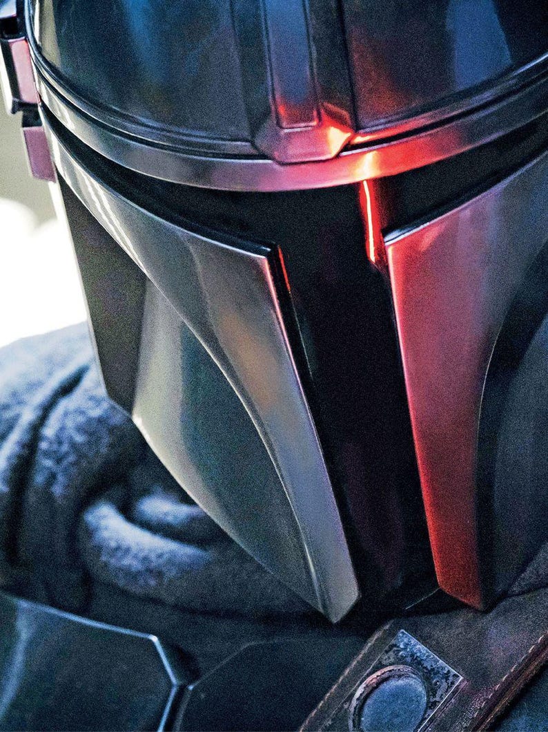 May include: Close-up of a Mandalorian helmet. The helmet is primarily dark gray with silver and red accents. The design features angular shapes and a visor with a red light. The helmet is part of a costume.