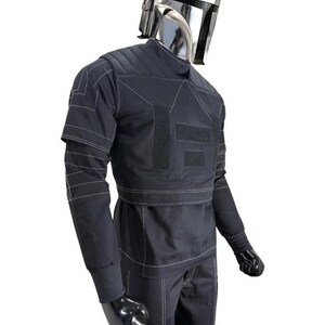 Op de afbeelding: Een zwart kostuum in tactische stijl met een metallic zilveren helm met een T-vormig vizier. De outfit bestaat uit een top met lange mouwen met witte contraststiksels, gewatteerde schouderpanelen en rechthoekige klittenbandsluitingen.