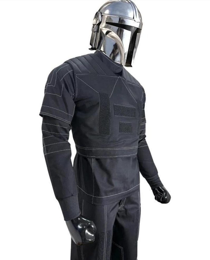 Op de afbeelding: Een zwart kostuum in tactische stijl met een metallic zilveren helm met een T-vormig vizier. De outfit bestaat uit een top met lange mouwen met witte contraststiksels, gewatteerde schouderpanelen en rechthoekige klittenbandsluitingen.