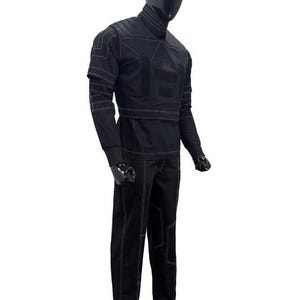 Op de afbeelding: Een zwarte set bestaande uit een shirt met lange mouwen en een bijpassende broek op een etalagepop. De outfit heeft een hoge kraag, gestructureerde schouderpanelen en opvallende witte contraststiksels die geometrische patronen vormen.