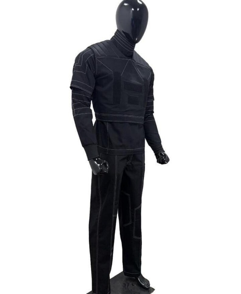 Op de afbeelding: Een zwarte set bestaande uit een shirt met lange mouwen en een bijpassende broek op een etalagepop. De outfit heeft een hoge kraag, gestructureerde schouderpanelen en opvallende witte contraststiksels die geometrische patronen vormen.
