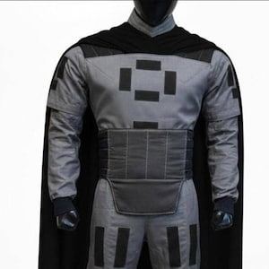 May include: A gray long-sleeved tunic and pants set featuring black rectangular patches on the chest, shoulders, and legs. The outfit includes a wide, horizontally stitched black waist belt and a long black cape attached at the shoulders.