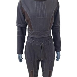 May include: A dark gray jumpsuit with white stitching details. The top features a high collar, long sleeves, and a fitted waist. The pants have a button closure and pockets. The jumpsuit has brown accents and is displayed on a mannequin.