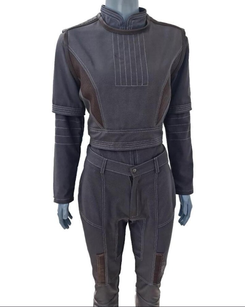 May include: A dark gray jumpsuit with white stitching details. The top features a high collar, long sleeves, and a fitted waist. The pants have a button closure and pockets. The jumpsuit has brown accents and is displayed on a mannequin.