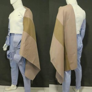 Puede incluir: Un maniqu&iacute; viste una parte superior blanca de cuello alto, pantalones cargo de color azul claro con bolsillos laterales y una capa de tela drapeada en dos tonos, beige y tostado. El conjunto se muestra de perfil y desde la parte trasera.