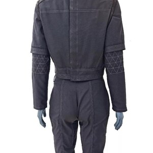 May include: A dark gray jumpsuit with white stitching details. The jacket has a stand-up collar and a cropped waist. The sleeves feature geometric patterns near the cuffs. The pants have a fitted design. The outfit is displayed on a mannequin.