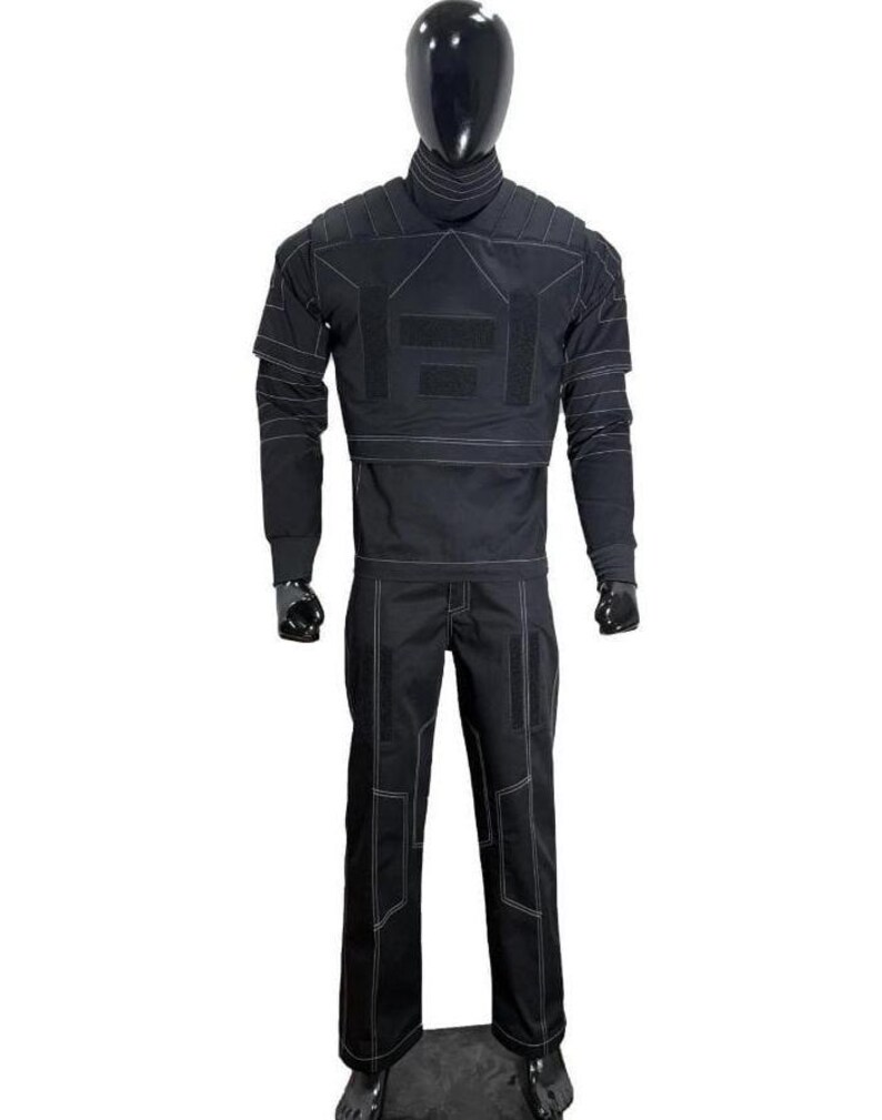 Op de afbeelding: Een zwarte tactische tweedelige outfit bestaande uit een top met lange mouwen met een hoge kraag en een bijpassende broek. Het kledingstuk is voorzien van opvallende witte contraststiksels, gestructureerde schouderpanelen en klittenband.