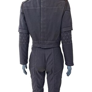 May include: Dark gray jumpsuit with white stitching details. The jacket has a cropped design with a belt, and the sleeves feature geometric patterns. The shoulders and collar have brown accents. The pants are fitted with visible seams.