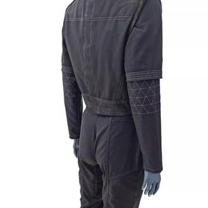May include: Dark gray jumpsuit with white stitching details. Features a stand-up collar, shoulder accents, and geometric patterns on the sleeves. The jumpsuit is shown on a mannequin.