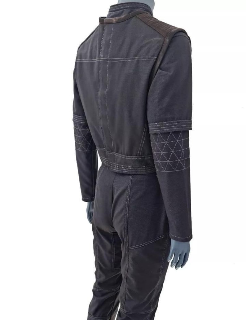 May include: Dark gray jumpsuit with white stitching details. Features a stand-up collar, shoulder accents, and geometric patterns on the sleeves. The jumpsuit is shown on a mannequin.