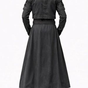 K&ouml;nnte beinhalten: Ein schwarzes Outfit bestehend aus einer cropped Jacke mit Schulterdetails, einem langen Rock und einer Hose. Die Jacke hat lange &Auml;rmel mit einem dezenten Streifenmuster. Der Rock ist weit und flie&szlig;end, und die Hose ist gerade geschnitten.