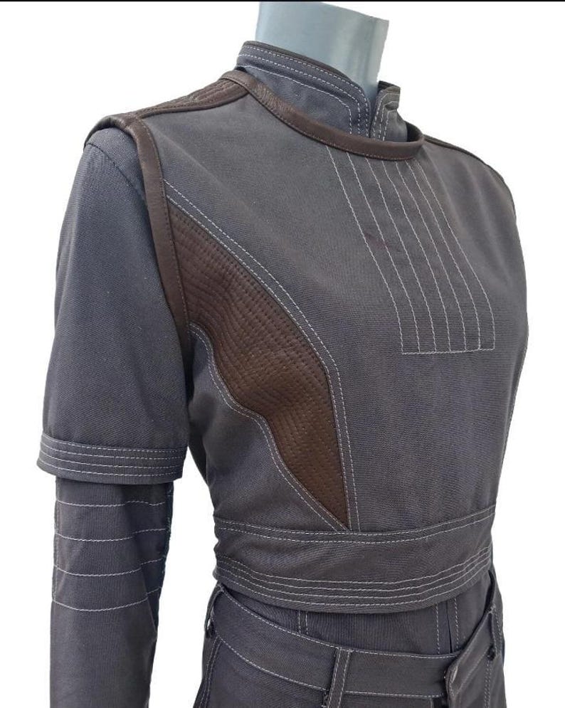 May include: A gray jumpsuit with brown accents and white stitching. The short-sleeved top features a high collar and a textured brown panel on the shoulder. The waist is cinched with a matching belt.