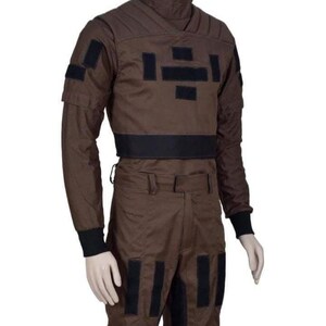 May include: Brown jumpsuit with black accents. The outfit features a high collar, long sleeves, and multiple black rectangular patches on the chest, arms, and legs. The waist is cinched with a wide black band, and the cuffs are black.
