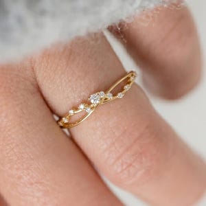 Puede incluir: Un anillo de oro delicado con un diseño de banda fina entrelazada. El anillo está adornado con una serie de pequeñas piedras claras de corte redondo engastadas a lo largo de los hilos de metal que se cruzan en el dedo.