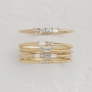Könnte beinhalten: Eine Sammlung zarter goldener Ringe, jeder mit kleinen, funkelnden Edelsteinen. Einige Ringe haben eine Reihe von drei rechteckigen Steinen, während andere eine Ansammlung kleinerer, runder Steine haben. Die Ringe werden auf einer hellen Oberfläche präsentiert.