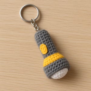 Puede incluir: Un llavero de ganchillo hecho a mano con forma de linterna. La linterna es gris con una banda amarilla y un botón amarillo. El extremo de la luz es blanco. Tiene un anillo de metal plateado.