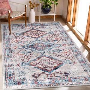 Puede incluir: Una alfombra rectangular con un diseño desgastado de inspiración vintage. La alfombra presenta un fondo gris claro con un medallón central y un borde en tonos de azul, naranja y rojo. La alfombra está colocada sobre un suelo de madera.