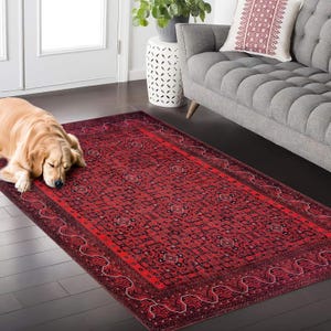 Könnte beinhalten: Ein rechteckiger roter Teppich mit detailliertem geometrischem Muster und dunklem Rand. Der Teppich liegt auf einem dunklen Holzboden, mit einem schlafenden Golden Retriever auf der linken Seite. Ein graues Sofa und ein weißer Beistelltisch sind im Hintergrund.