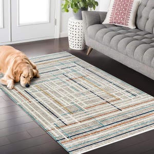 Könnte beinhalten: Ein rechteckiger Teppich mit einem geometrischen Muster in Beige, Weiß, Blau und Orange. Der Teppich hat einen Rand und ein gitterartiges Design. Ein Golden Retriever liegt auf dem Teppich.