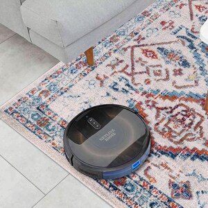 May include: A black robotic vacuum cleaner on a patterned rug. The vacuum is round with a low profile and the text "NETLINE HOME" is visible. The rug has a cream background with blue and orange geometric designs. A gray sofa and white table are in the background.