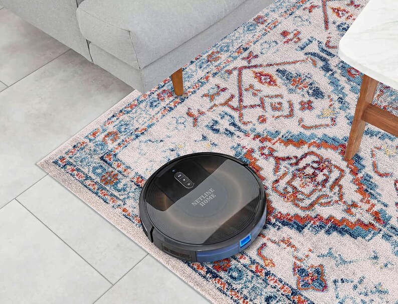 May include: A black robotic vacuum cleaner on a patterned rug. The vacuum is round with a low profile and the text "NETLINE HOME" is visible. The rug has a cream background with blue and orange geometric designs. A gray sofa and white table are in the background.