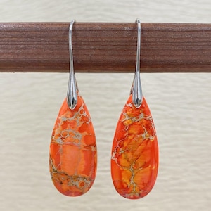 Peut inclure: Une paire de boucles d'oreilles en forme de goutte d'eau avec un motif marbré orange et jaune vif. Les boucles d'oreilles ont des crochets argentés et sont présentées sur une tige en bois. Bijoux.