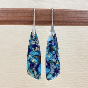 May include: A pair of dangle earrings with a silver hook and a teardrop-shaped stone. The stone features a mosaic pattern of blue, teal, and brown hues. The earrings are displayed against a wooden background.