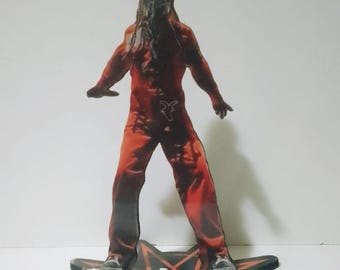 May include: A red and black Slipknot band member figure. The figure is wearing a red jumpsuit and has long, dark hair. The figure stands on a black star-shaped base with the band's logo in white.