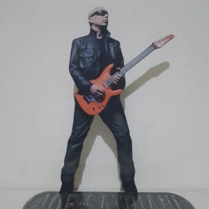 May include: A figurine of Joe Satriani, a guitarist, wearing sunglasses and a black leather jacket, holding an orange electric guitar. The figurine stands on a gray base with the name "JOE SATRIANI" engraved on it.
