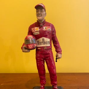 Pode incluir: Estatueta de um piloto de corrida com um fato vermelho, segurando um capacete. O fato tem logotipos de patrocinadores, incluindo "Marlboro". A estatueta está sobre uma base preta com o nome "Michael Schumacher."