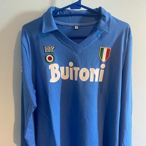 May include: A light blue long-sleeve shirt with a V-neck collar. The shirt features the word "Buitoni" in white block letters across the chest, along with two patches. One patch has the letters "NR" and the other has the colors of the Italian flag.