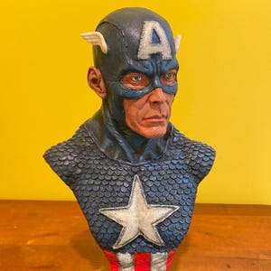 Puede incluir: Busto detallado de Capit&aacute;n Am&eacute;rica, con un casco azul con una "A" blanca y alas, una m&aacute;scara azul y un pecho estrellado. La escultura est&aacute; montada sobre una base negra, mostrando la imagen del ic&oacute;nico superh&eacute;roe.