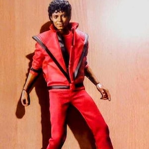 May include: A figurine of Michael Jackson in a red outfit, reminiscent of his Thriller era. The figure is posed in a dance stance, with the word "Thriller" on the base. The outfit includes a red jacket with black accents and matching pants.